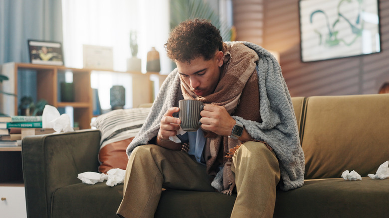 A young man sits on the couch wrapped in blankets and blowing on a hot drink
