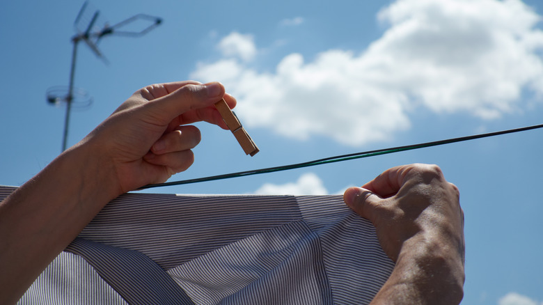 A pair of hands holding a wooden clothespin and a shirt about to be hung on a clothing line outside.