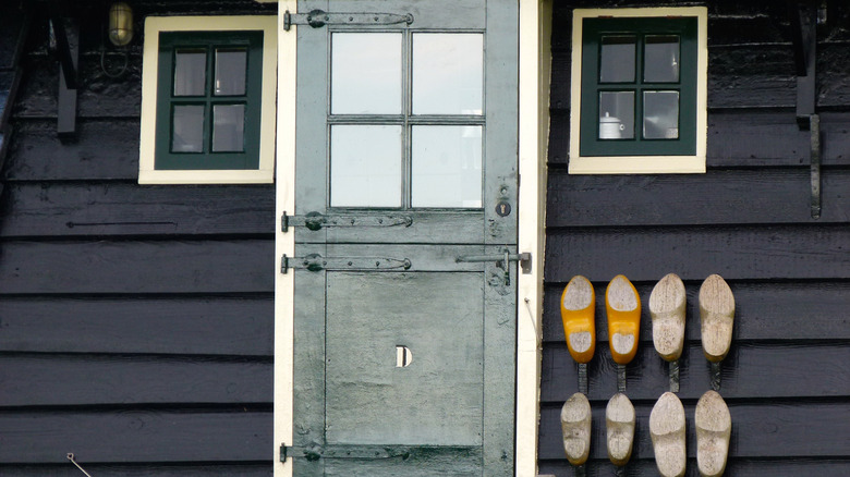 A gray Dutch door is flanked by two windows and several pairs of wooden clogs
