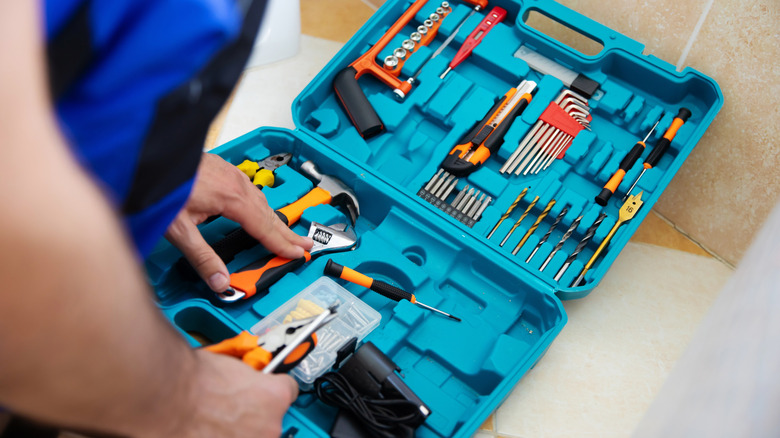 A person's hands reaching inside an open blue plastic toolbox with orange hand tools inside