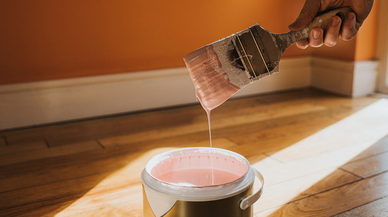 A hand holding a paint brush with pink paint dripping off into paint can and orange wall behind