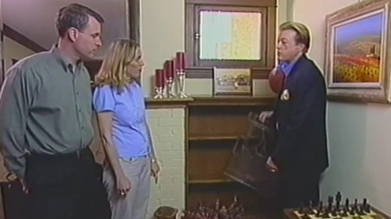 Three people standing in a living space next to a brown shelf.