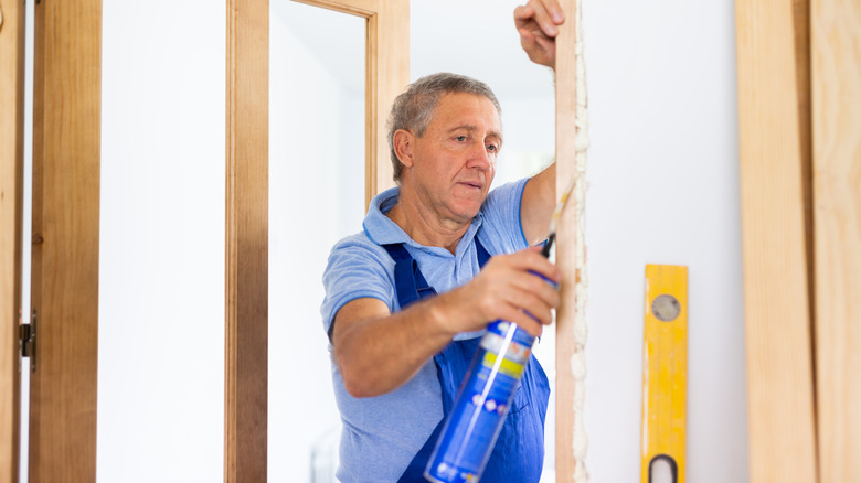 Middle-aged contractor in a blue uniform installing foam sealant in door jamb.