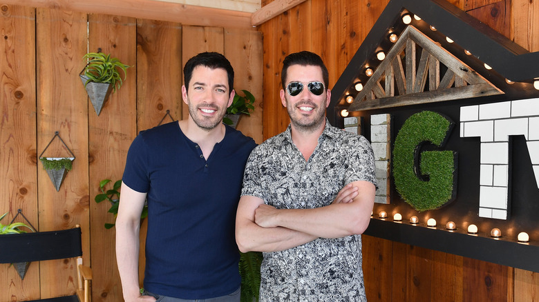 Property Brothers Jonathan and Drew Scott posing against rustic backdrop with HGTV sign.