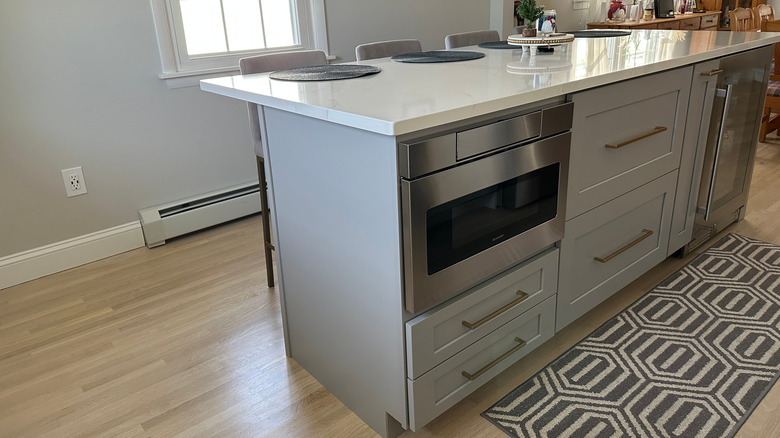 Modern kitchen island with built-in stainless steel microwave drawer, shaker-style cabinets with brass hardware, and a white quartz countertop