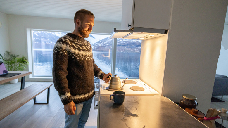 A man makes a hot drink and wears a sweater inside a cold kitchen.
