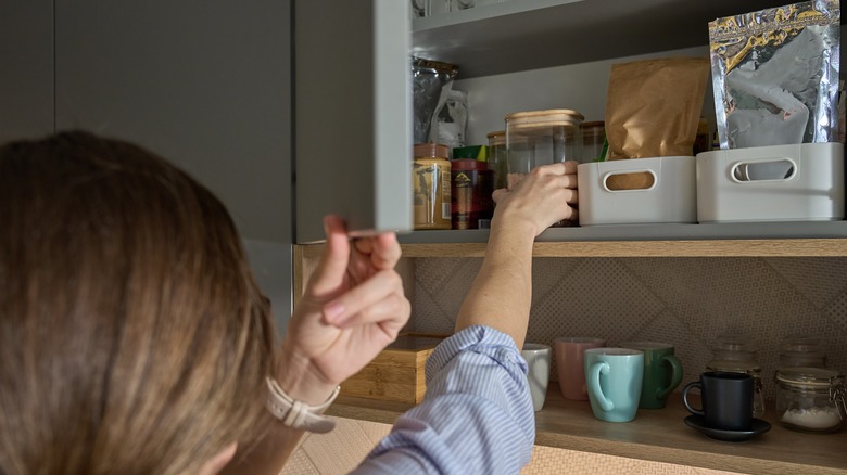 A person takes a glass jar from a kitchen cabinet with food containers and glassware inside.