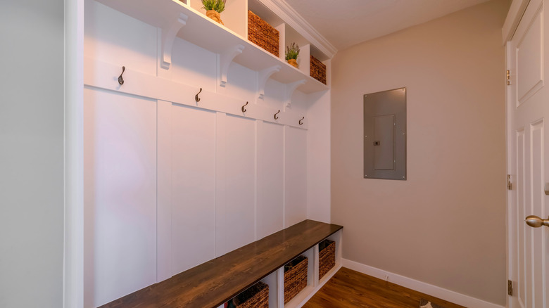 Mudroom interior with white wall and dark wood floor and seat. There is a white door on the left across the storage baskets below and above the seat with white wood panel with hooks at the back.