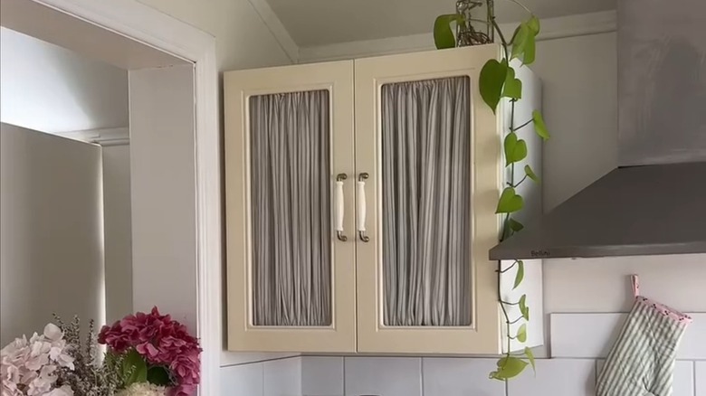Kitchen cabinet with glass doors, simple neutral striped fabric curtains inside concealing contents.