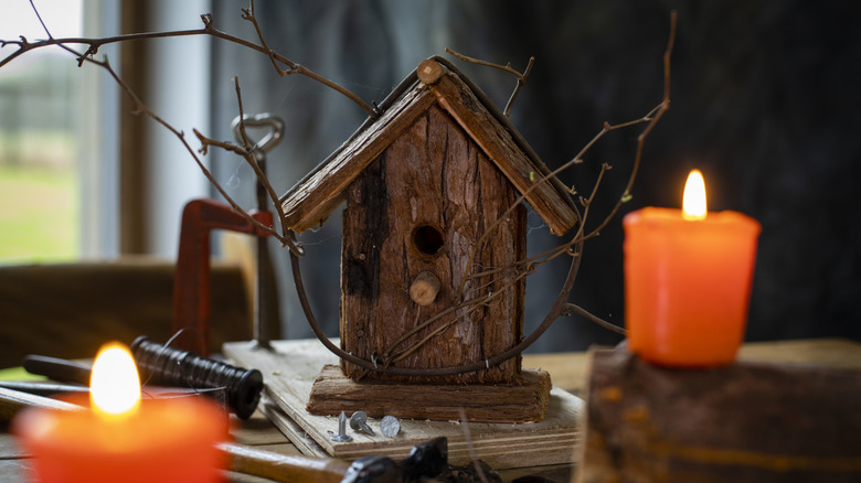 A birdhouse sits on a rustic table between two candles