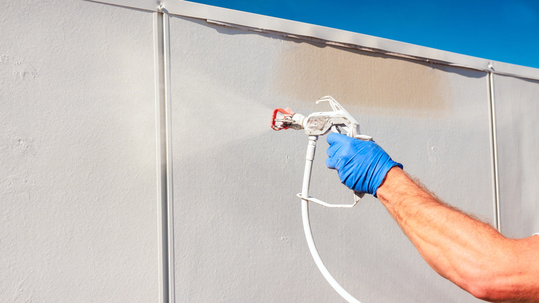 Close-up of a blue-gloved hand using a paint sprayer to coat a wall