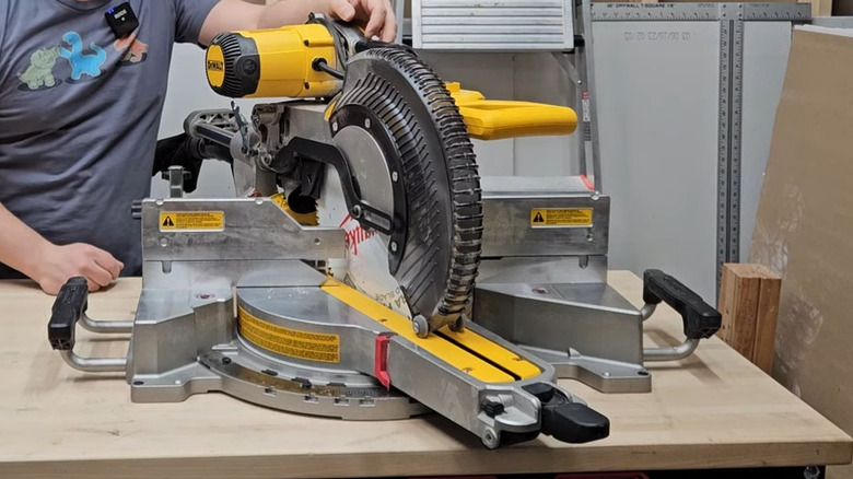 Man in dinosaur shirt standing behind a DeWalt compound miter saw in a workshop or garage