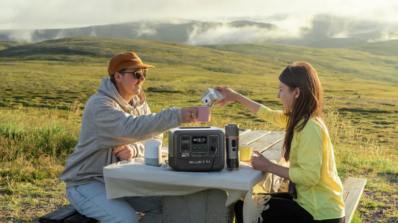 a Bluetti power station sitting on a table between two campers who are in a field drinking coffee