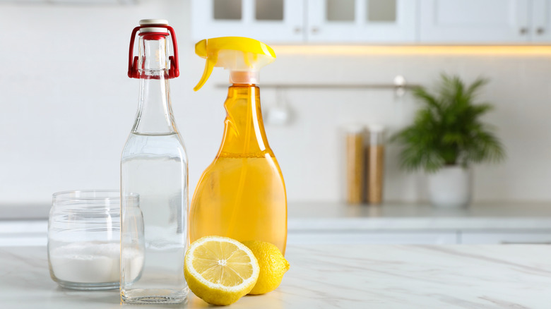A yellow spray bottle, a cut lemon, a bottle of vinegar, and jar of baking soda rest on a clean kitchen counter.