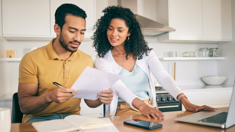 A couple going over paperwork and financial documents at kitchen table with laptop and smartphone next to them