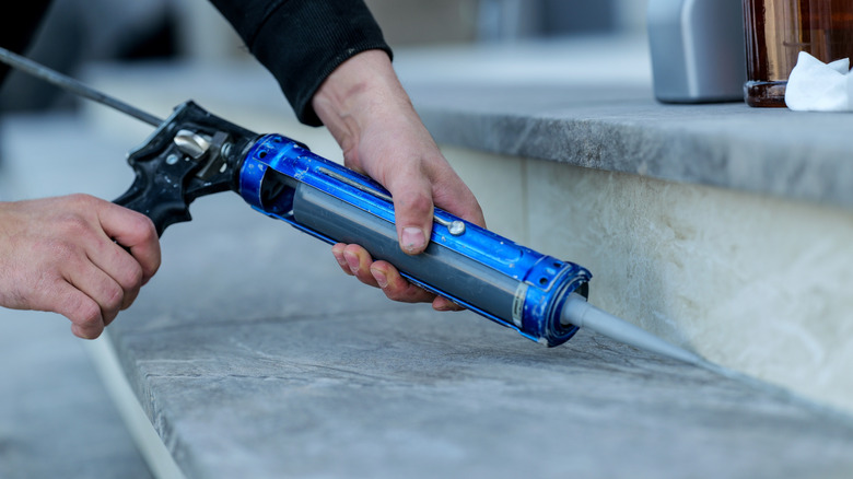 Person's hands applying gray caulk to stone tile steps outdoors.