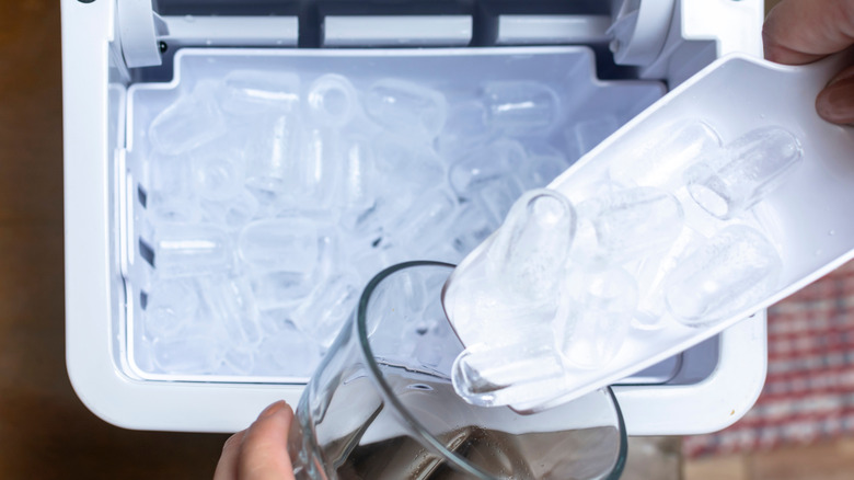 A person using a white scoop to get ice from a stand alone ice maker and putting it into a glass of water