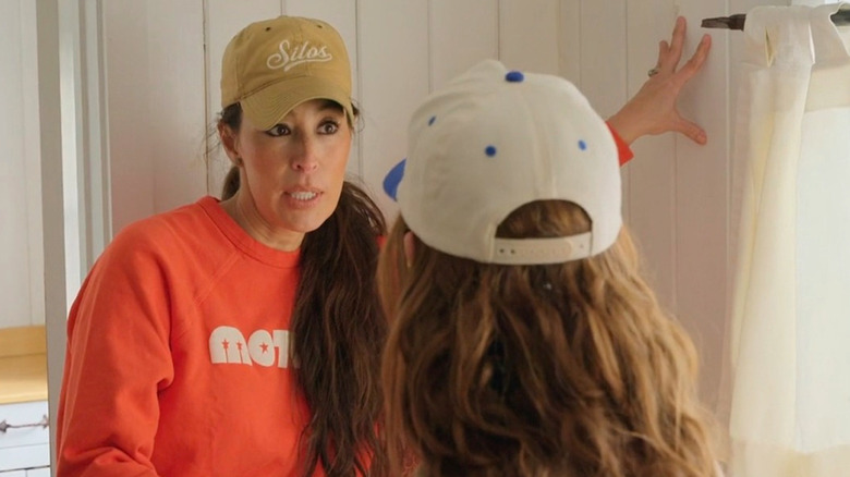 Joanna Gaines wearing an orange sweatshirt and putting hand on wall talking to another woman whose back is to camera