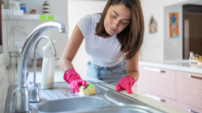 A woman is wearing pink gloves and scrubbing around a kitchen sink