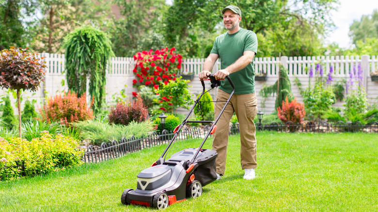 A person mowing the lawn in a small backyard with flower beds and a fence.
