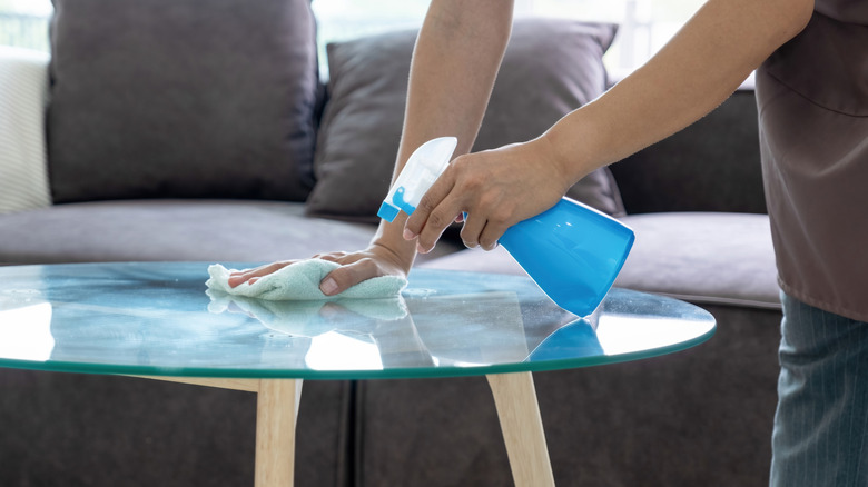 A housekeeper in an apron holds spray bottle of rubbing alcohol and a cloth while cleaning a glass table in a bright, tidy living room at home.