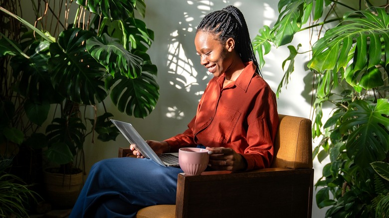 A smiling person holds a cup of coffee while looking down at a laptop in a plant-filled room