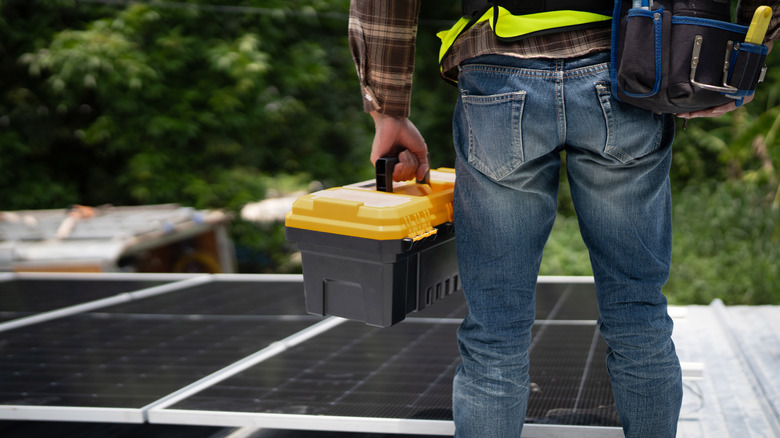 A solar panel installer stands on a rooftop holding a yellow toolbox