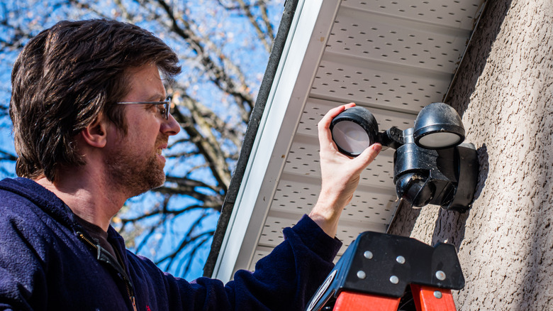 A man in a blue sweater and glasses looking at black motion sensor lights installed outside a home