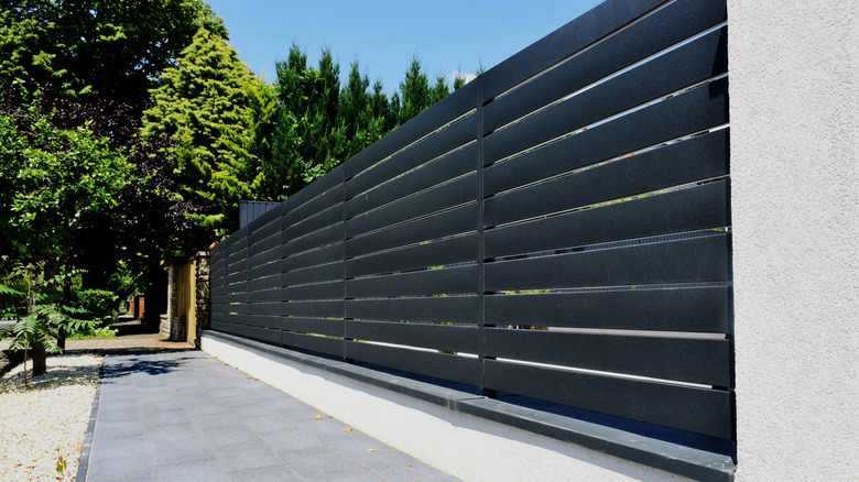 A tall fence made of horizontal aluminum slats next to a tree-lined sidewalk