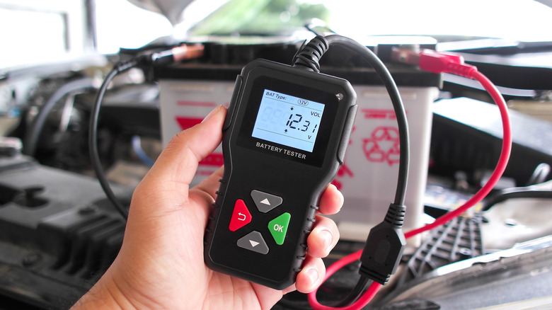 A digital battery tester displays a 12.31V reading while connected to a 12V lead-acid battery.