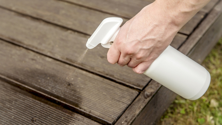 Hand spraying white bottle onto wooden deck outdoors.