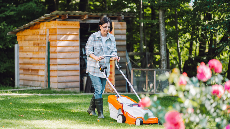 A woman pushes an orange electrical mower over grass in front of a wooden shed
