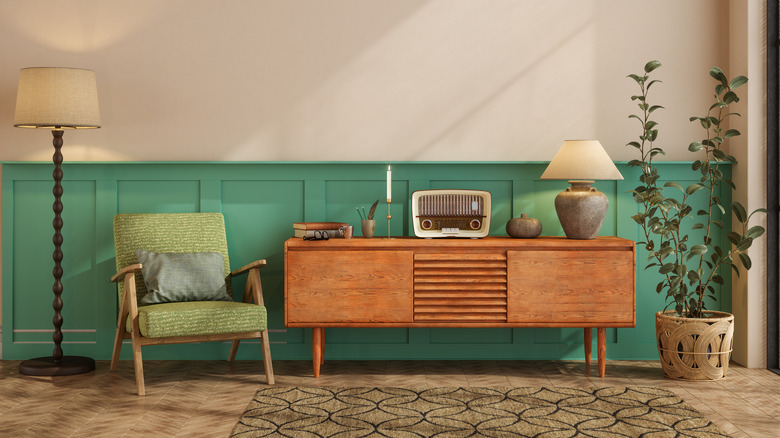 Wooden console table with vintage radio, lamp, books, and candle on top, next to a green armchair, standing lamp, and tall plant