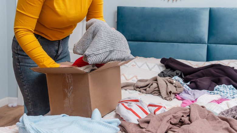 A person sorting clothes that are laid out on bed and putting them into a cardboard box