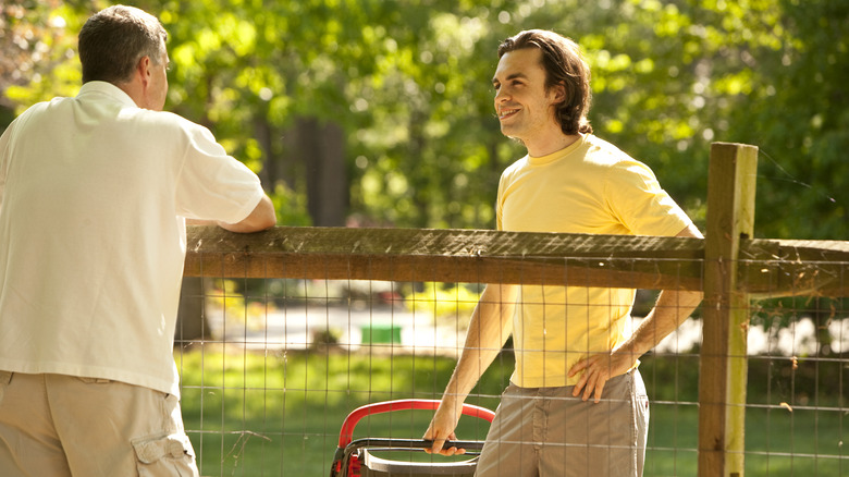 A man approaches his neighbor who is mowing his lawn.