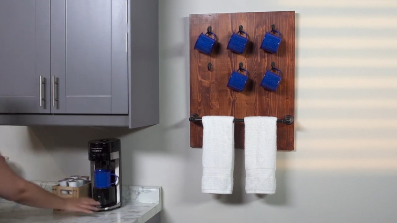A wooden coffee mug rack fitted with a towel rail and blue cups hangs on the wall in a kitchen.