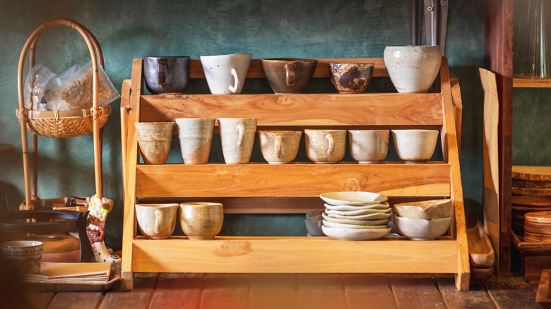 Handmade ceramic cups and plates sit on a wooden countertop spice rack in a kitchen.