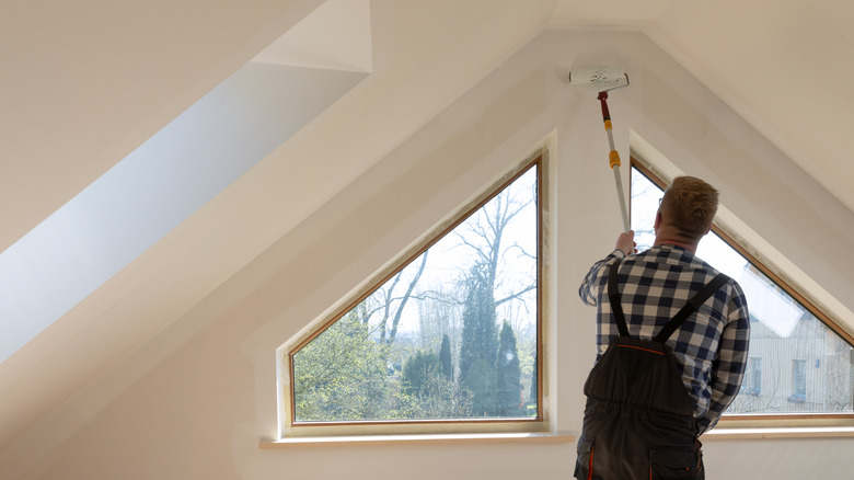 A handyman uses an extended roller to paint attic walls above a set of windows
