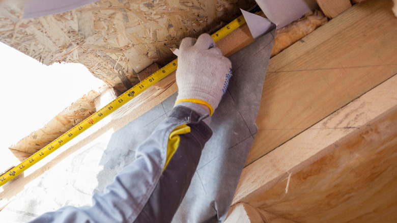 A person measures a sloped roof in an attic with a measuring tape
