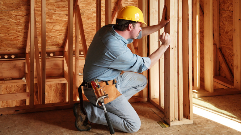 A man wearing a yellow hardhat marks a measurement on a stud wall