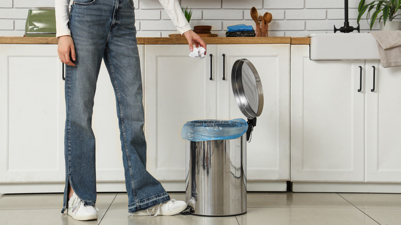 Woman throwing crumpled paper into trash bin with foot pedal in kitchen.