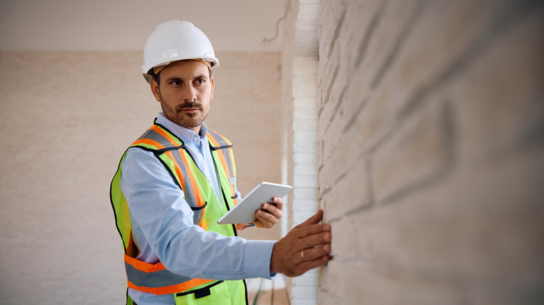 Man in hard hat and yellow vest inspecting a brick wall in an apartment holding a tablet