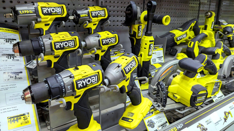 Display of Ryobi 18V ONE+ power tools on a pegboard wall, showcasing cordless drills, impact drivers, and other yellow and black battery-powered tools in a hardware store setting.