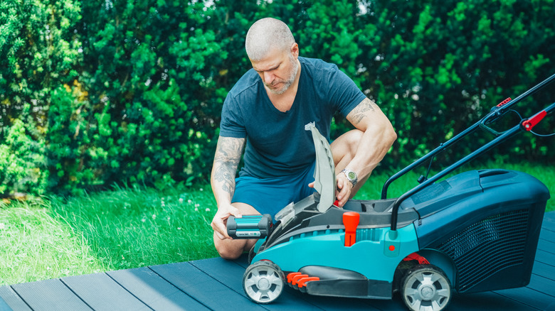A man in casual summer clothes installing a battery into a modern electric lawn mower while kneeling on a wooden deck in a lush green garden.