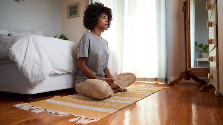 A woman sitting at the food of the bed on a rug in a meditative pose