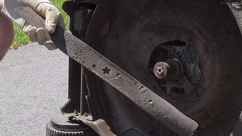 A person's gloved hand holding a replacement mower blade in front of a push mower on its side