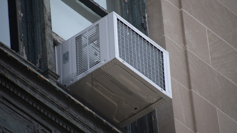 An air conditioner unit juts out of a wooden windowsill on a gray brick building.