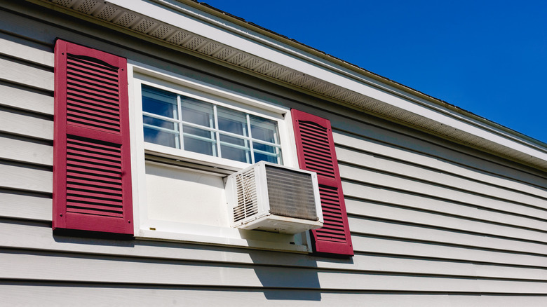 An AC unit sits in a residential window that features red shutters.