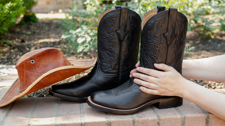 A person holding black cowboy boots with brown hat