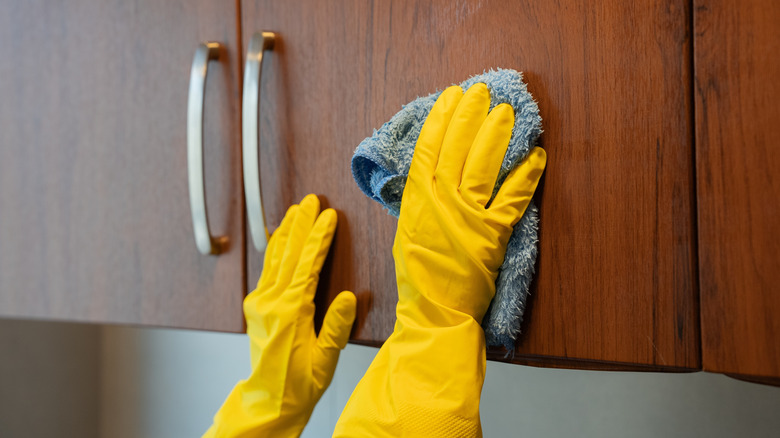 Yellow-gloved hands wipe wooden kitchen cabinets with blue cloth.